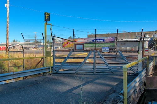 A secured gate covered with barbed wire, marked as Gate F, blocks a road leading to an industrial facility. Various warning signs are displayed, including a 'No Trespassing' notice. The background features industrial buildings and clear blue skies.