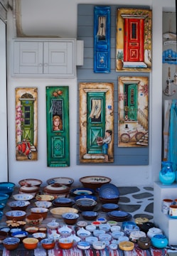 An array of colorful, hand-painted ceramic bowls and plates laid on a patterned rug. Behind them, a wall displays decorative paintings of doors, each uniquely colorful and detailed, with elements such as a bicycle, a child holding a pot, and a vintage scooter.
