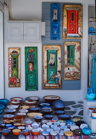 An array of colorful, hand-painted ceramic bowls and plates laid on a patterned rug. Behind them, a wall displays decorative paintings of doors, each uniquely colorful and detailed, with elements such as a bicycle, a child holding a pot, and a vintage scooter.