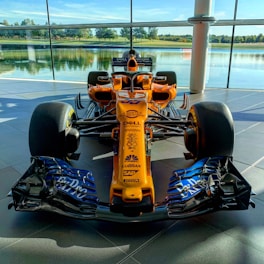 A brightly colored Formula 1 racing car is displayed indoors, set against large glass windows with a serene outdoor view of a lake and trees. The car's orange body features various sponsor logos in contrasting colors. The sleek design includes prominent aerodynamic features, with large front and rear wings and exposed tires.