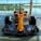 A brightly colored Formula 1 racing car is displayed indoors, set against large glass windows with a serene outdoor view of a lake and trees. The car's orange body features various sponsor logos in contrasting colors. The sleek design includes prominent aerodynamic features, with large front and rear wings and exposed tires.