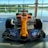 A brightly colored Formula 1 racing car is displayed indoors, set against large glass windows with a serene outdoor view of a lake and trees. The car's orange body features various sponsor logos in contrasting colors. The sleek design includes prominent aerodynamic features, with large front and rear wings and exposed tires.