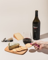A bottle of red wine labeled 'ONDULÉ NAPA VALLEY CALIFORNIA 2013' is positioned next to a glass of wine and a selection of cheeses and crackers on a wooden board. A person's hand with pink nail polish is holding a cracker. A corkscrew and cork are visible, both branded with 'ONDULÉ'. The setting suggests a casual wine and cheese tasting.