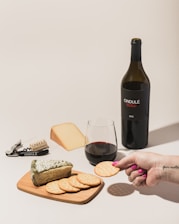 A bottle of red wine labeled 'ONDULÉ NAPA VALLEY CALIFORNIA 2013' is positioned next to a glass of wine and a selection of cheeses and crackers on a wooden board. A person's hand with pink nail polish is holding a cracker. A corkscrew and cork are visible, both branded with 'ONDULÉ'. The setting suggests a casual wine and cheese tasting.