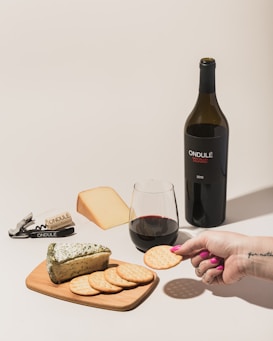 A bottle of red wine labeled 'ONDULÉ NAPA VALLEY CALIFORNIA 2013' is positioned next to a glass of wine and a selection of cheeses and crackers on a wooden board. A person's hand with pink nail polish is holding a cracker. A corkscrew and cork are visible, both branded with 'ONDULÉ'. The setting suggests a casual wine and cheese tasting.