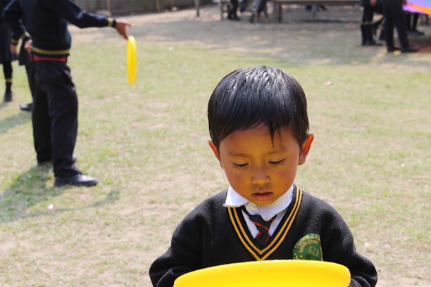 A young child wearing a school uniform stands on a grassy field while holding a yellow plastic disc. Other children in similar uniforms can be seen in the background, suggesting a playground or school environment.