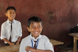 Two smiling children wearing school uniforms stand in a classroom. Both are dressed in white shirts and ties. One child is in the foreground, clearly delighted, while the other is slightly further back, with a similar joyful expression. Wooden desks and a brown wall are visible in the background.