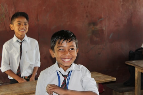 Two smiling children wearing school uniforms stand in a classroom. Both are dressed in white shirts and ties. One child is in the foreground, clearly delighted, while the other is slightly further back, with a similar joyful expression. Wooden desks and a brown wall are visible in the background.