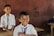 Two smiling children wearing school uniforms stand in a classroom. Both are dressed in white shirts and ties. One child is in the foreground, clearly delighted, while the other is slightly further back, with a similar joyful expression. Wooden desks and a brown wall are visible in the background.
