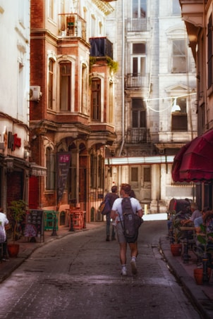 A narrow cobblestone street flanked by old, ornate buildings with rustic facades. People stroll down the path, including an individual wearing a backpack, suggesting a casual or tourist-like atmosphere. Potted plants and small tables line the sidewalks outside cafes, adding a quaint charm to the scene. The light filters gently through the urban landscape, enhancing the warmth of the red and brown tones in the architecture.