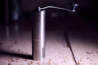 A metallic manual coffee grinder with a crank handle is placed on a worn, rustic surface. The grinder's body is cylindrical with a small viewing window and appears to be made of stainless steel. The scene is dimly lit, producing subtle reflections on the metal surface.