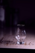 A clear, empty whiskey glass sits on a reflective surface, which appears to be slightly stained or textured. The background is dark and out of focus, emphasizing the glass.