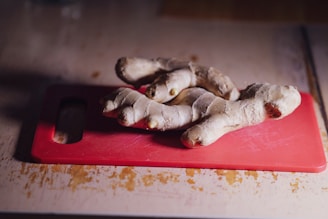 A piece of fresh ginger root is placed on a red cutting board, which is set on a light-colored wooden surface. The ginger shows a robust texture with knobby sections and a natural, brownish skin tone.