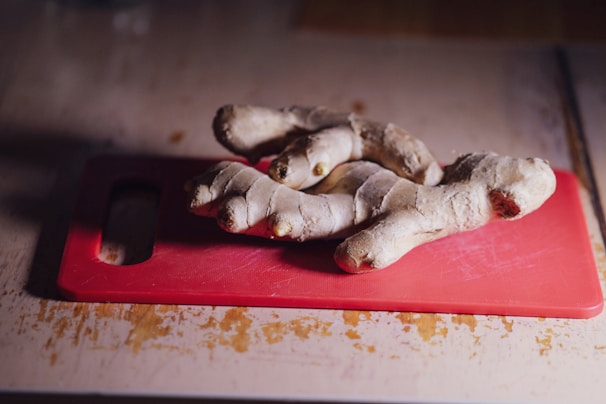 A piece of fresh ginger root is placed on a red cutting board, which is set on a light-colored wooden surface. The ginger shows a robust texture with knobby sections and a natural, brownish skin tone.