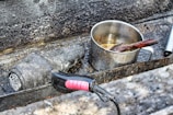 A rusted and weathered stovetop features a well-used metal pot with visible burn marks and a wooden spoon inside. Beside it, a dirty and charred hair dryer lies on the surface, both items suggesting abandonment or extensive use.