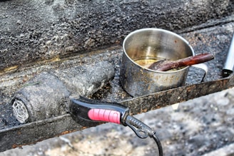 A rusted and weathered stovetop features a well-used metal pot with visible burn marks and a wooden spoon inside. Beside it, a dirty and charred hair dryer lies on the surface, both items suggesting abandonment or extensive use.
