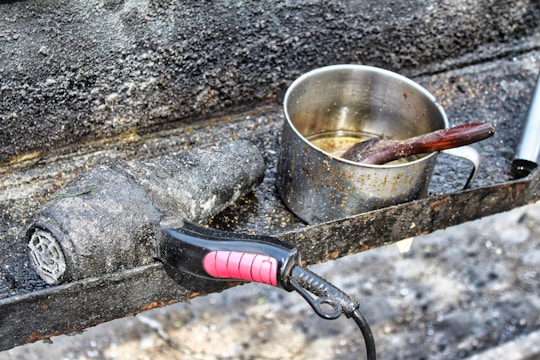 A rusted and weathered stovetop features a well-used metal pot with visible burn marks and a wooden spoon inside. Beside it, a dirty and charred hair dryer lies on the surface, both items suggesting abandonment or extensive use.