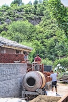 Construction workers are actively building a brick structure. One person is operating a cement mixer while others are arranging bricks. The construction site is set against a backdrop of lush green vegetation and a rocky hill, indicating a rural or suburban environment.
