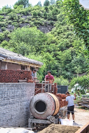Construction workers are actively building a brick structure. One person is operating a cement mixer while others are arranging bricks. The construction site is set against a backdrop of lush green vegetation and a rocky hill, indicating a rural or suburban environment.