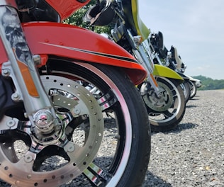 A row of motorcycles lined up on a gravel surface, showing close-up views of their wheels and front fenders. The motorcycles display various colors including red and green, with chrome details and intricate designs visible on the front forks.