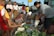 A bustling street market scene with several people engaged in buying and selling produce. Various fresh vegetables and fruits are arranged on the ground, with sellers and buyers interacting closely. A woman in a grey shirt is packaging items, while another person hands over money. A few others are standing around observing or waiting their turn.