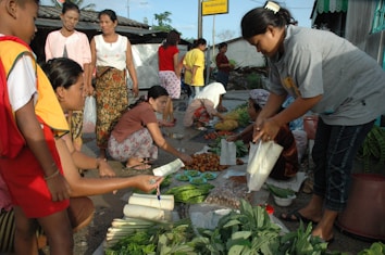 A bustling street market scene with several people engaged in buying and selling produce. Various fresh vegetables and fruits are arranged on the ground, with sellers and buyers interacting closely. A woman in a grey shirt is packaging items, while another person hands over money. A few others are standing around observing or waiting their turn.