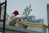 A person is painting intricate and colorful patterns on a structure. The design features swirling motifs with vibrant accents in blue, red, green, and yellow. The individual is wearing a white shirt and a red cloth covering their head.