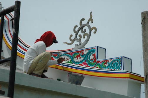 A person is painting intricate and colorful patterns on a structure. The design features swirling motifs with vibrant accents in blue, red, green, and yellow. The individual is wearing a white shirt and a red cloth covering their head.
