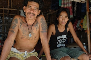 Two men sit together in a shaded, bamboo-structured setting. One man is shirtless, showcasing several tattoos and a pendant necklace. The other man wears a black tank top with a graphic print. Hanging clothes and fabrics are visible in the background, adding a sense of casual, everyday life.