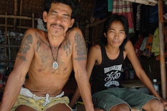 Two men sit together in a shaded, bamboo-structured setting. One man is shirtless, showcasing several tattoos and a pendant necklace. The other man wears a black tank top with a graphic print. Hanging clothes and fabrics are visible in the background, adding a sense of casual, everyday life.