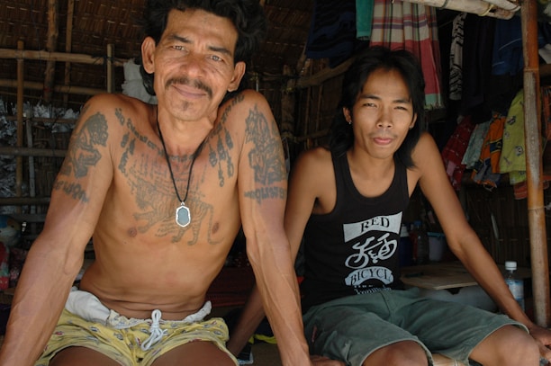 Two men sit together in a shaded, bamboo-structured setting. One man is shirtless, showcasing several tattoos and a pendant necklace. The other man wears a black tank top with a graphic print. Hanging clothes and fabrics are visible in the background, adding a sense of casual, everyday life.