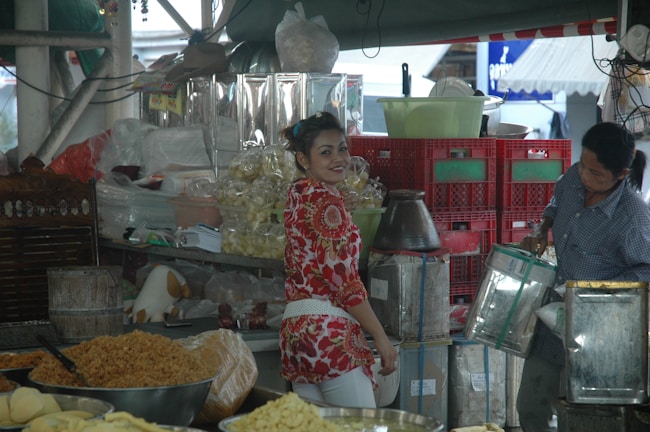A bustling market scene featuring two people. One person in a red and white floral shirt is smiling, standing at a vibrant stall filled with a variety of goods, including packaged snacks and grains. Another person is handling a metal container, surrounded by stacks of red crates and bags of produce.