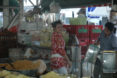 A bustling market scene featuring two people. One person in a red and white floral shirt is smiling, standing at a vibrant stall filled with a variety of goods, including packaged snacks and grains. Another person is handling a metal container, surrounded by stacks of red crates and bags of produce.