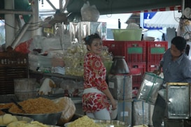 A bustling market scene featuring two people. One person in a red and white floral shirt is smiling, standing at a vibrant stall filled with a variety of goods, including packaged snacks and grains. Another person is handling a metal container, surrounded by stacks of red crates and bags of produce.