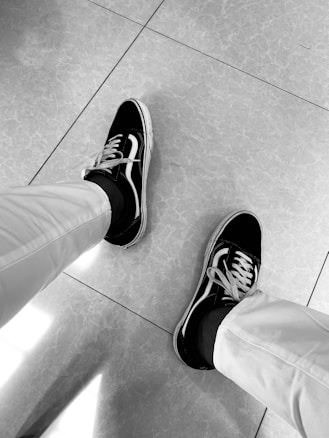 A pair of feet wearing black and white canvas sneakers with white laces, and dressed in light-colored pants. The person is standing on a tiled floor that has a light pattern.