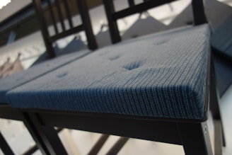 A modern chair with a textured blue cushion and dark wooden frame is captured from a low angle. The cushion features subtle button tufting, adding a touch of sophistication. The background is blurred, highlighting the chair as the focal point.