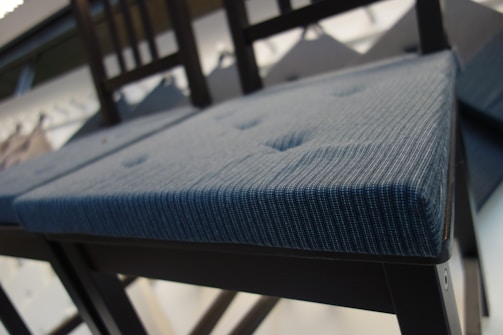 A modern chair with a textured blue cushion and dark wooden frame is captured from a low angle. The cushion features subtle button tufting, adding a touch of sophistication. The background is blurred, highlighting the chair as the focal point.
