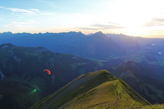 A scenic mountain landscape with a grassy peak. A single paraglider with a red canopy floats in the air, set against a backdrop of distant mountain ranges under a clear sky. The sun is setting on the horizon, casting a warm glow over the scene.
