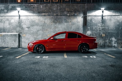 A parked red car is situated in a dimly lit urban parking lot. The vehicle is in a well-marked parking space against a concrete wall, which is somewhat illuminated by two overhead street lamps casting soft shadows. The environment gives a slightly gritty, urban vibe with visible parking space numbers painted on the ground and wall.