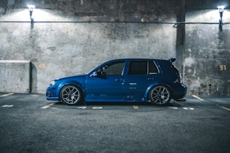 A blue car is parked in an illuminated parking garage with a concrete wall backdrop. The car is positioned in an empty parking space, surrounded by numbered parking spots 48, 49, and 51. The scene has minimal lighting from two overhead fixtures, creating a moody atmosphere.