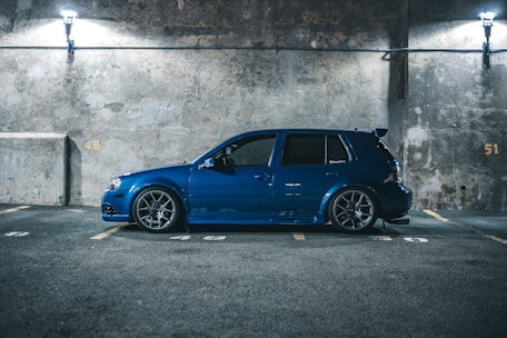 A blue car is parked in an illuminated parking garage with a concrete wall backdrop. The car is positioned in an empty parking space, surrounded by numbered parking spots 48, 49, and 51. The scene has minimal lighting from two overhead fixtures, creating a moody atmosphere.