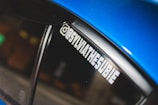 A close-up of a car window featuring a decal with the text '@SYLVIATHESUBIE' in bold white letters. The car appears to be a vibrant blue color with a glossy finish.
