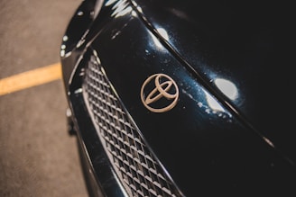 Close-up of a car's front, focusing on the Toyota emblem placed on a dark, glossy surface with a mesh grille below. The car surface reflects light, showcasing its sleek finish.