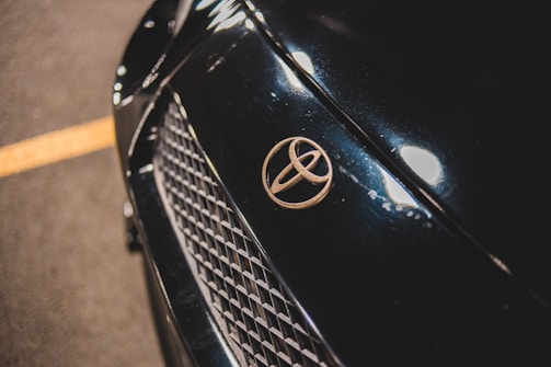 Close-up of a car's front, focusing on the Toyota emblem placed on a dark, glossy surface with a mesh grille below. The car surface reflects light, showcasing its sleek finish.