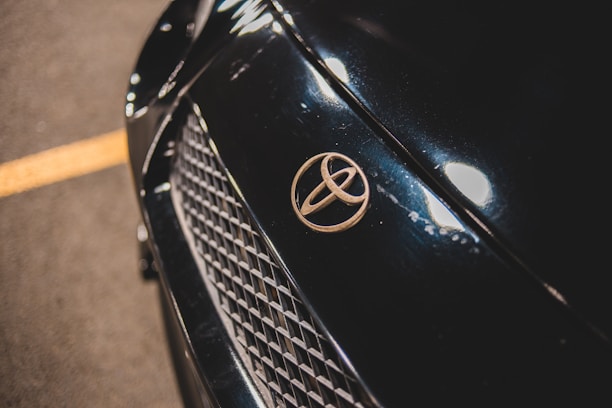 Close-up of a car's front, focusing on the Toyota emblem placed on a dark, glossy surface with a mesh grille below. The car surface reflects light, showcasing its sleek finish.