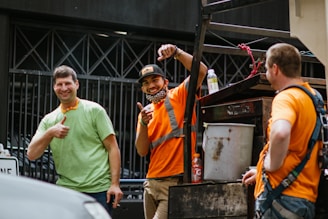 Three men wearing construction attire are cheerfully interacting near a construction site. One man in a green shirt is giving a thumbs-up, while another in an orange shirt with a reflective vest is pointing and smiling. The third man is wearing an orange shirt as well, facing away. A metal scaffold or piece of machinery is in the background, with various items like a bottle and a bucket placed on it.