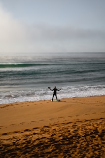A person in a wetsuit stands on a sandy beach holding a paddle, gazing towards the ocean. The waves are gently crashing onto the shore under a cloudy sky. Footprints are visible in the sand, suggesting recent movement.
