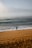 A person in a wetsuit stands on a sandy beach holding a paddle, gazing towards the ocean. The waves are gently crashing onto the shore under a cloudy sky. Footprints are visible in the sand, suggesting recent movement.