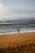 A person in a wetsuit stands on a sandy beach holding a paddle, gazing towards the ocean. The waves are gently crashing onto the shore under a cloudy sky. Footprints are visible in the sand, suggesting recent movement.