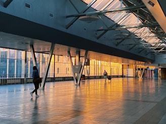 A spacious corridor with large windows showcasing a warm glow from the sunset. Two individuals walk across the shiny tiled floor, one carrying a backpack and the other pulling a suitcase. The architecture features high ceilings with exposed beams and angled support columns.